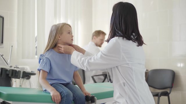 Tracking shot of female pediatrician in white coat checking tonsils or lymph nodes and throat of smiling little girl sitting on couch in doctors office, male colleague working at desk in background