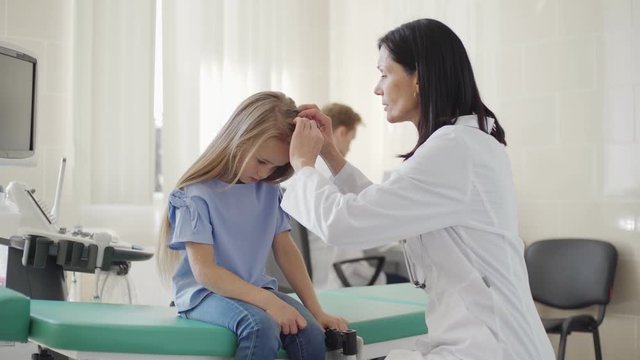 Tracking medium shot of female pediatrician in white coat checking blonde hair of little girl sitting on couch in doctors office, male doctor working at desk in background