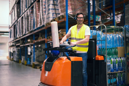 Professional warehouse worker with headset communication equipment driving forklift and relocating packages in storage center. Working at warehouse.