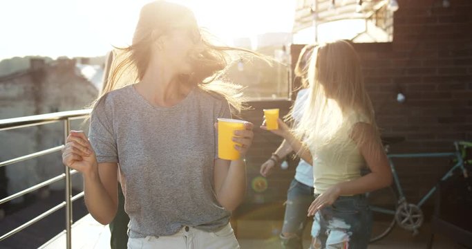 Attractive happy woman in casual outfit and glasses dancing on rooftop party, holding cup in hands, party with friends