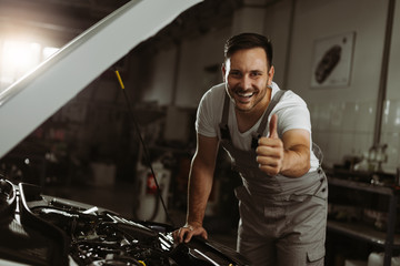 Happy mechanic showing thumbs up while working on a car in auto repair shop.He is looking at camera