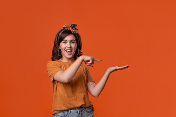 Beautiful smiling brunette woman in a t-shirt and headband holding palm of a hand for product placement standing isolated over orange background. Place for advertisement.