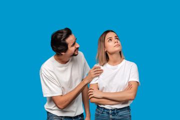 Brunet bearded man with mustaches in white t-shirt and blue jeans asking his girlfriend for forgiveness isolated over blue background. Concept of guilt.