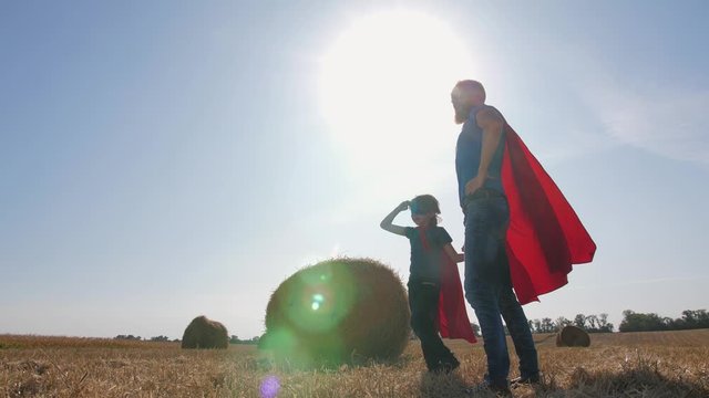 Playful readhead dad and son dressed as superheroes. Family looking into distance and stratching hands forward while standing on mowed wheat field against background of straw stacks and bright sundown