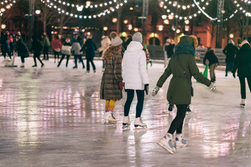 Group of girs skating back to us. Girlfriends ice skating in city park, snowy evening. Healthy outdoor winter activity