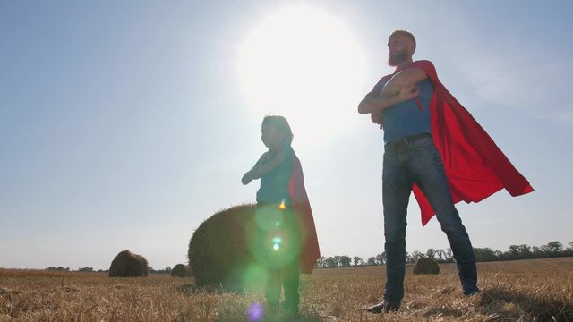 Bearded father and cute little son in superhero costumes standing in confident poses with hands on hips and arms folded. Family looking into distance on background of wheat straw stacks at sunset