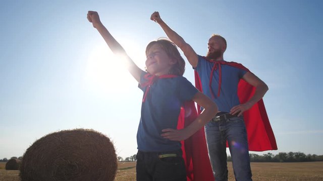 Confident father and son in superhero pose standing with hands on hips and looking into distance during sunset. Redhead man and child stretching forward clenched fist and saying superhero's motto