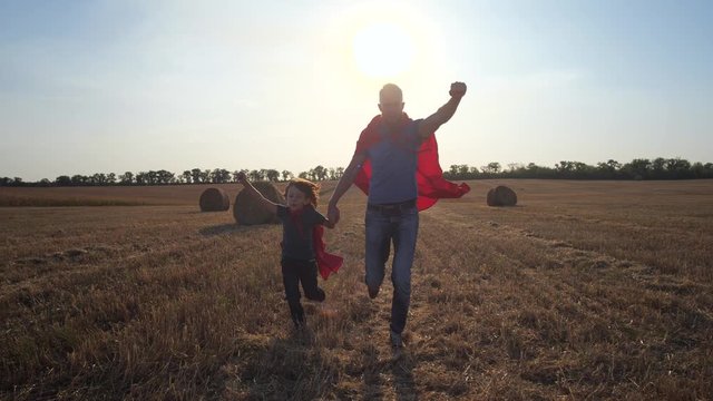 Playful dad and son in superhero costumes putting forward clenched fist running to rescue on background of sunset. Happy united family playing in superman capes running holding hands across field