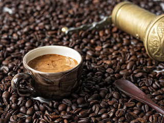 A black vintage cup with freshly brewed espresso stands in coffee beans. Nearby lies a manual bronze coffee grinder. Coffee grains are scattered on a wooden table. Close-up. Selective focus