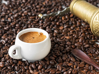 A white cup with freshly brewed espresso stands in coffee beans. Nearby lies a manual bronze coffee grinder. Coffee grains are scattered on a wooden table. Close-up. Selective focus