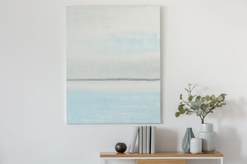 Pastel blue and white abstract oil painting on empty white wall with console table with flowers in vase and books