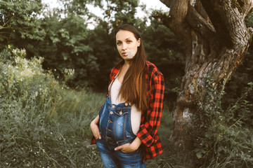 Beautiful caucasian pregnant woman in denim overalls with a flower in her mouth outside.