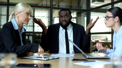 Black man showing no idea gesture listening to arguing female colleagues