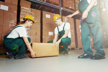 Two young warehouse workers in dark green uniforms and yellow helmets lifting heavy package