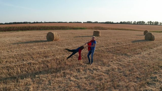 Drone shot of joyful family in costumes of superheroes having fun on wheat field with straw stacks. Aerial view of loving father whirling son while playing supermen
