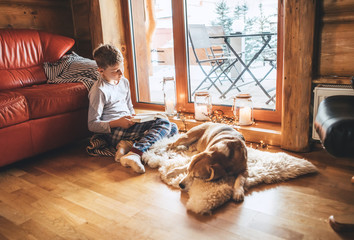 Boy reading book on the floor near slipping his beagle dog on sheepskin in cozy home atmosphere. Peaceful moments of cozy home concept image.
