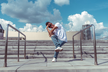 young guy a dancer in a white T-shirt, jeans, dancing break dance in summer in the city, in sunglasses, white sneakers background building clouds, active hip hop, youth lifestyle