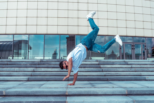 young guy a dancer in a white T-shirt, jeans, dancing break dance in summer in the city, in sunglasses, white sneakers background building clouds, active hip hop, youth lifestyle