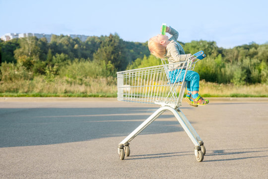 child boy blond in blue pants sits in a metal grocery trolley from a supermarket drinks juice throwing his head on an asphalt surface in the summer against a background of green plants