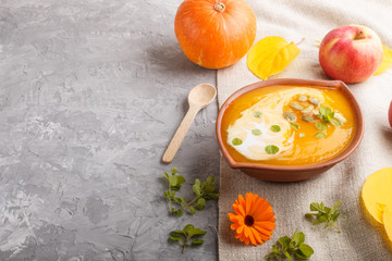 Traditional pumpkin cream soup with seeds in clay bowl on a gray concrete background with linen textile. side view