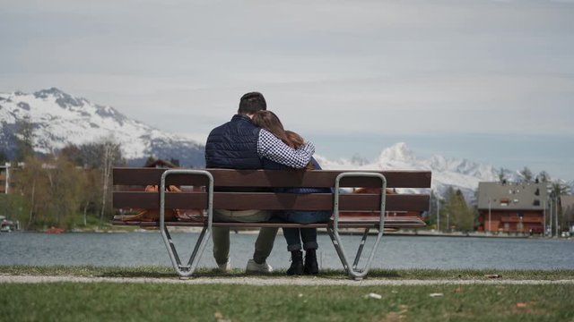 Young couple cuddling on lakeside wooden bench with beautiful mountain view. Rear view