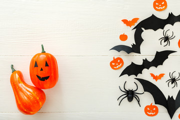 Halloween background with jack-o'-lanter, pumpkins, paper bats, spiders, confetti on white wooden background. Halloween holiday decorations. Flat lay, top view. Party invitation mockup, celebration.
