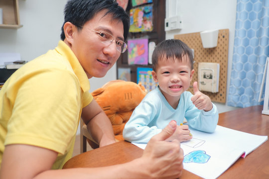 Asian dad and son showing thumbs up after finishing homework, Cute 3 - 4 years old toddler boy kid painting with crayons, Dad spending quality time with son after work, photo in real life interior