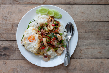 Top view of fried basil leave with squid and fried egg on rice in white plate with spoon and fork on wooden table.