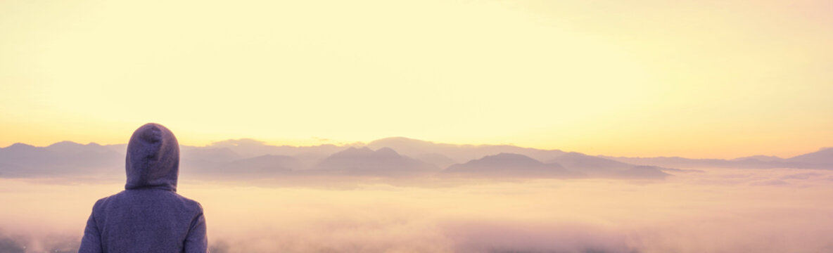 Woman with hood jacket cover head standing behind the fench on the top of the mountain looking out misty in the valley early morning twilight sky before sunrise sky