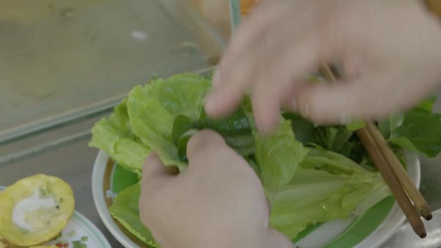 A daylight extreme closeup shot of a customer wrapping a mini rice pancake in a leaf of green lettuce, dipping it in a chili vinegar sauce and then taking a bite out of it.