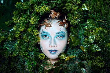 Woman fantasy make up with frogs on face and cover by green grass and leafs