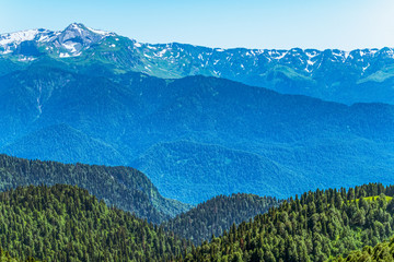Green mountain ridges, surrounded by high mountains. Snow-capped mountain peaks on the horizon. Krasnaya Polyana, Sochi, Caucasus, Russia.