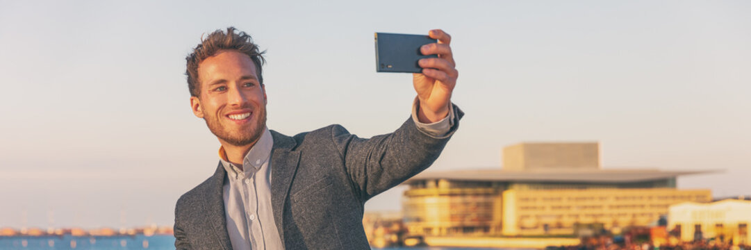 Phone using businessman taking selfie with mobile phone on travel business trip outside walking in city banner panorama. Young caucasian adult in his 30s. - Powered by Adobe