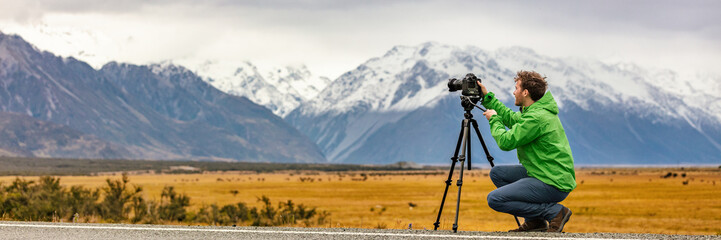 Photographer man taking nature photography with professional SLR camera at mountain landscape, New Zealand Banner. Tourist on travel adventure holiday shooting video on tripod panorama. - obrazy, fototapety, plakaty
