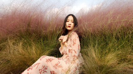 Portrait of a beautiful Chinese young woman in pink dress sitting in pink hairawn muhly field,...