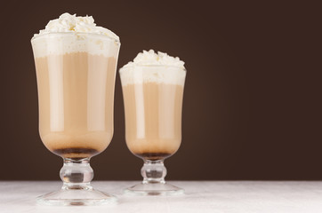 Two hot coffee drinks with whipped cream in wineglass in dark brown interior on white wooden table, copy space.