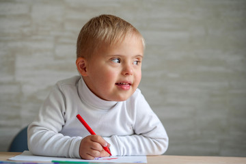 A little boy with Down syndrome sits at a wooden table and draws with a felt-tip pen on a cheat...
