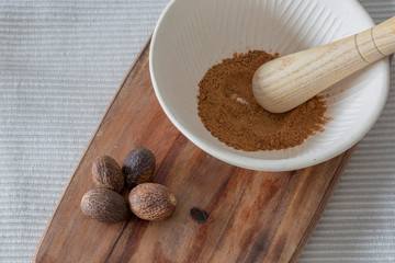 Ground nutmeg in a pestle and mortar with whole seeds on a wooden chopping board.  On a cream place mat.