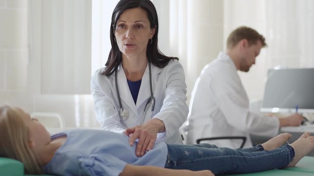 Tracking medium shot of female doctor in white coat palpating belly of girl lying on couch and talking to little patient, male doctor making notes in medical history at desk in background