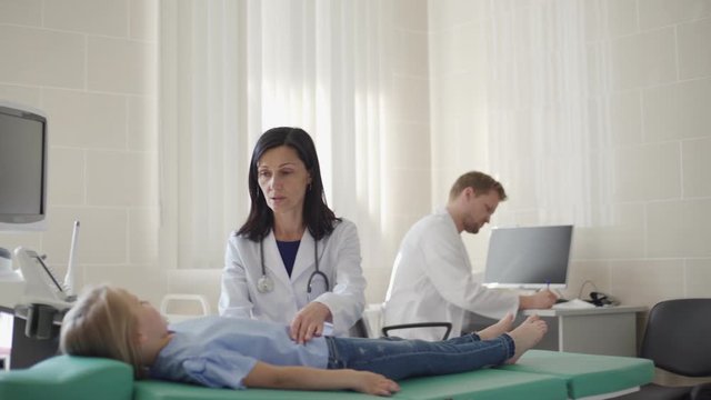Tracking medium shot of female doctor in white coat palpating belly of little girl lying on couch, male doctor making notes in medical history at desk in background