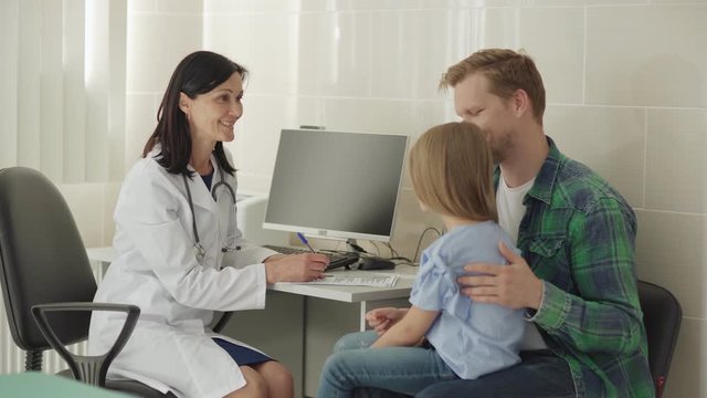 Middle aged female doctor in white coat sitting at desk in office and smiling while talking to father stroking gently back of little daughter sitting on his lap