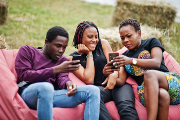 Three african american friends chill, sitting on poufs and using their phones outdoor.