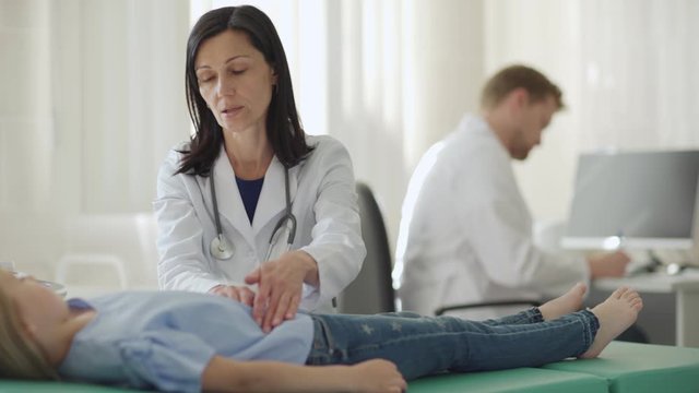 Tracking medium shot of female doctor in white coat palpating belly of little girl lying on couch and talking to male nurse making notes in medical history at desk in background