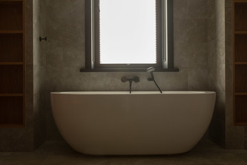 Fototapeta na wymiar White oval solid base freestanding bathtub with chrome mixer tap and hand shower mounted on wall installed under window