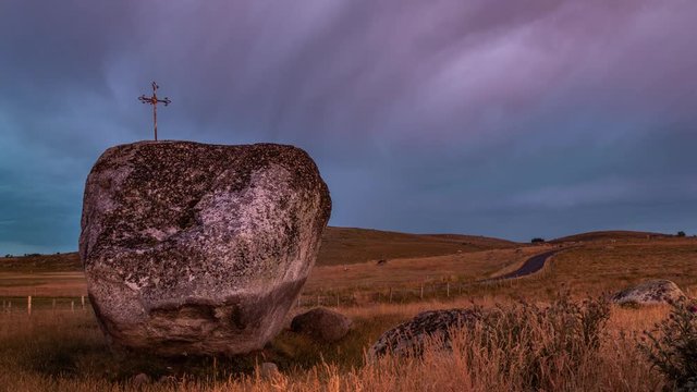 Time-lapse of a double rainbow during sunset with a small catholic cross planted in a boulder in the foreground, surrounded by peaceful hills and grass fields. Loz&egrave;re, Aubrac, France.