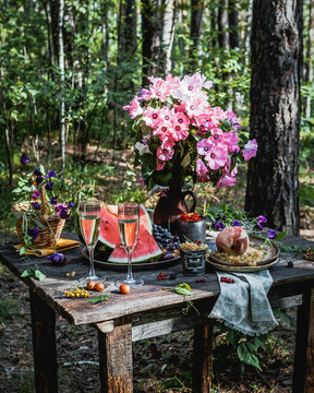 fresh berries in various mugs and plates, peach, watermelon slices and grape on wooden table with glasses of champagne and bouquet of pink flowers