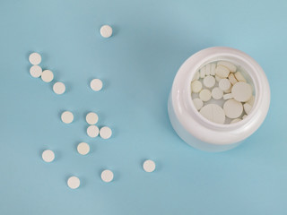 A bunch of white pills lying next to a discarded jar with pills on a blue background. Top view.