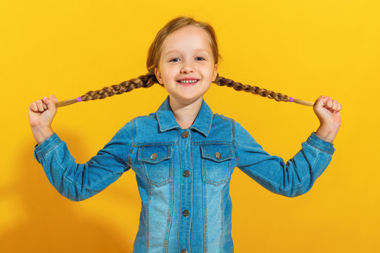 Portrait of a cheerful little girl in a denim shirt on a yellow background. The child holds pigtails of hair with his hands and looks at the camera