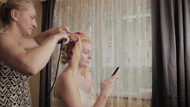 Professional hairdresser woman doing a hair-style photo of a model who is sitting with a phone in her hands.