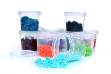 Open transparent plastic container with colorful plastic snap fastener buttons spilling out with other filled containers in background